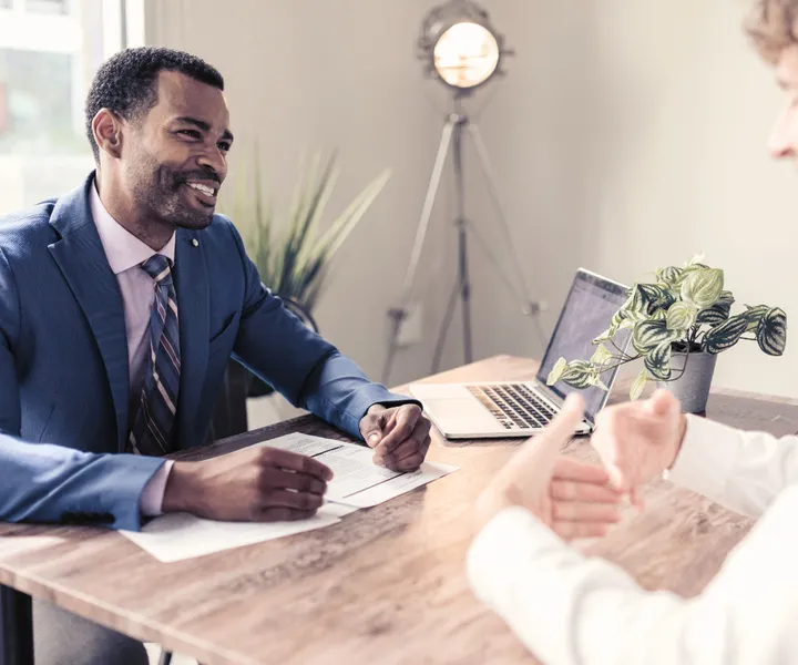 Two men sitting at a table in an office, engaged in conversation. One man wears a blue suit, and a laptop and plant are on the table.